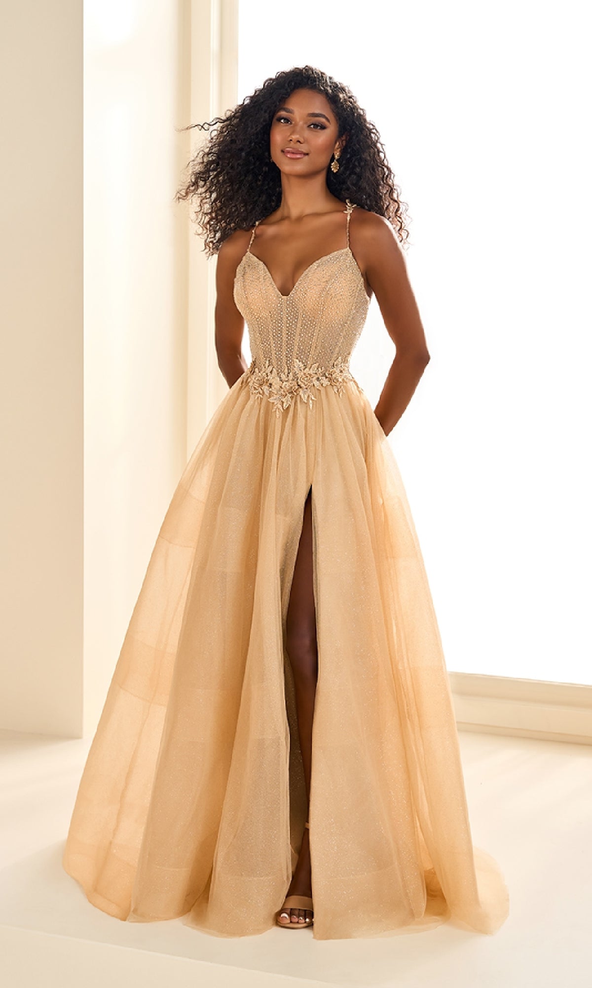 A woman stands in a bright room wearing the Ellie Wilde EW36058 long prom dress, featuring a light gold hue, corset bodice with spaghetti straps, floral waist details, high slit, and elegant floral lace embroidery.